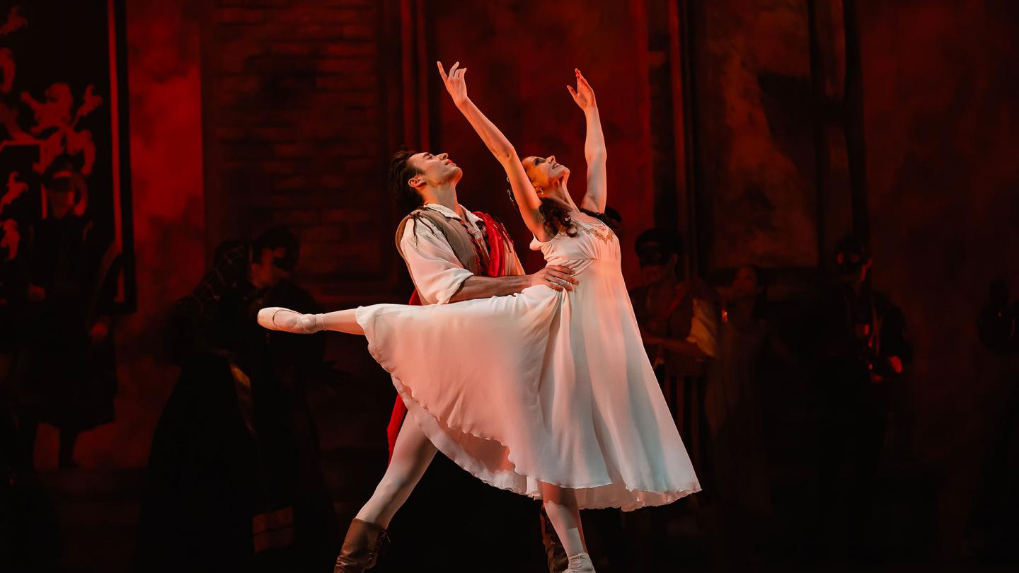 Juliet stands en pointe with her arms above her head, Romeo holds her by the waist