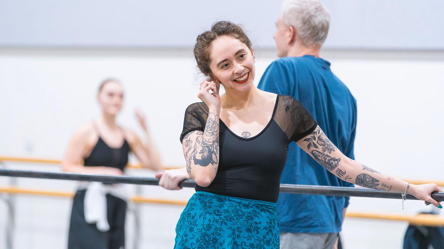 Woman in black top and blue slip stands at the barre, smiling
