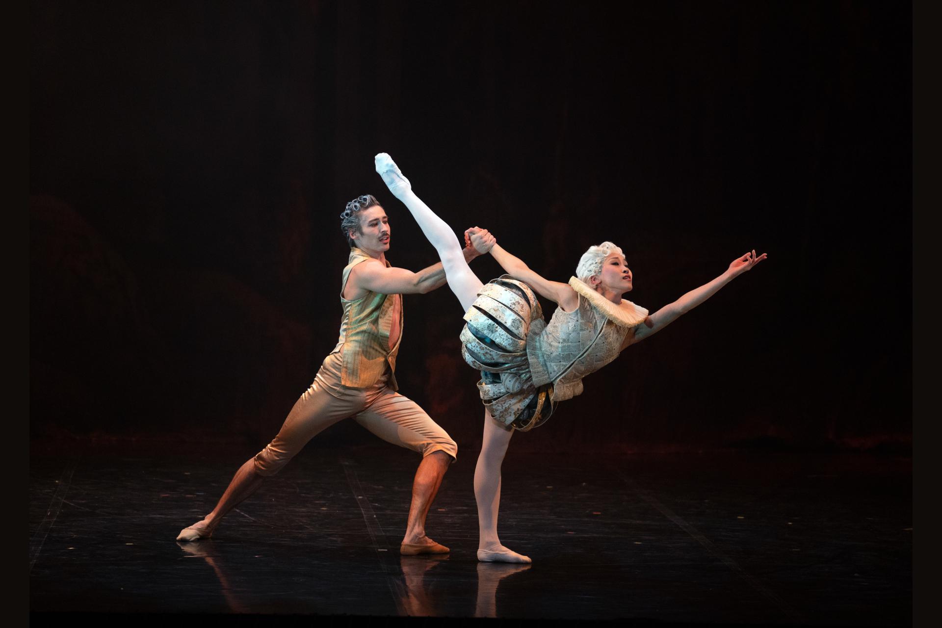 A female dancer in light 18th century costume reaches into the distance with her leg aloft, supported from behind by a male dancer in gold costume.