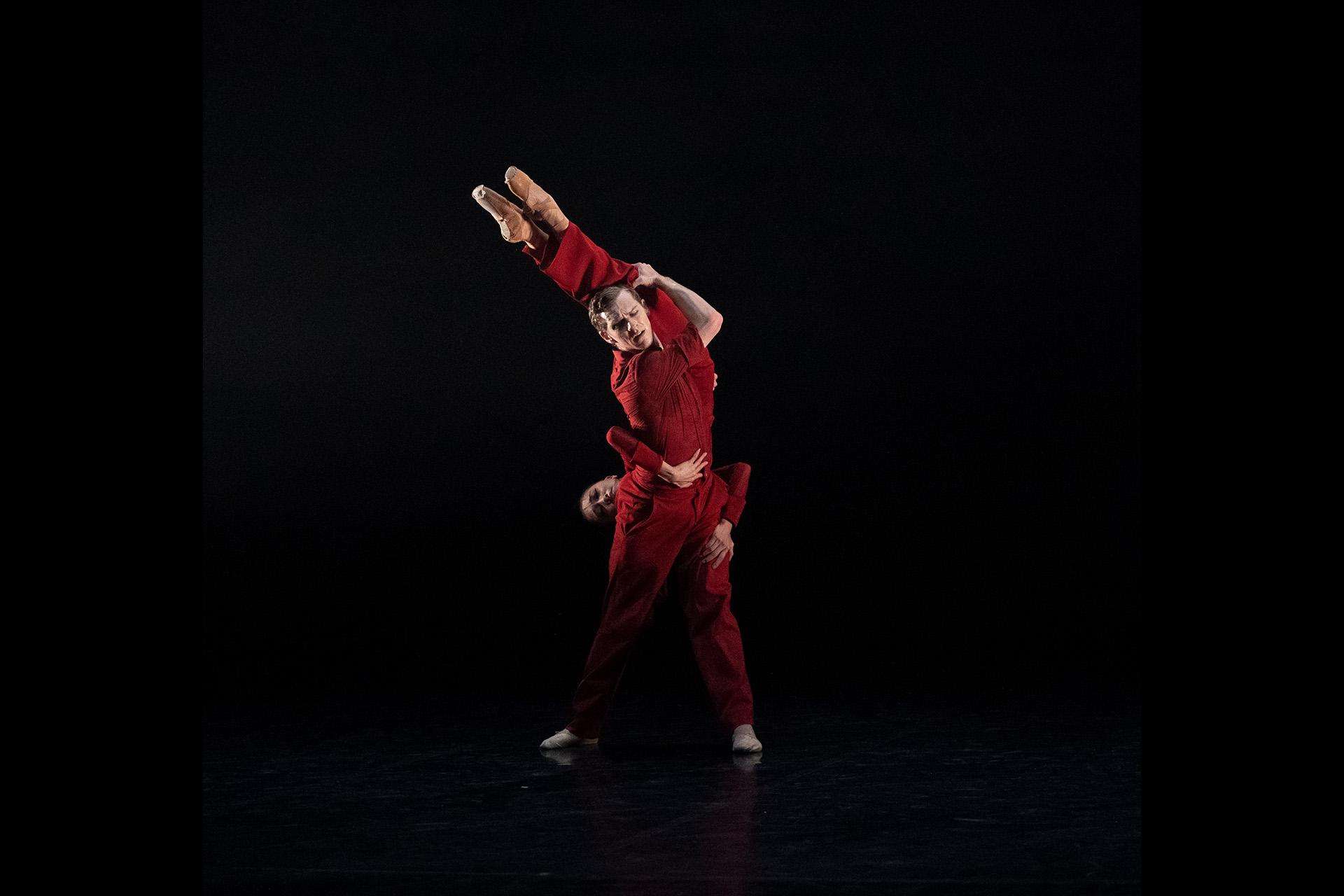 Two dancers dressed in red, the male holds the female with her feet together above his head and arms around his waist
