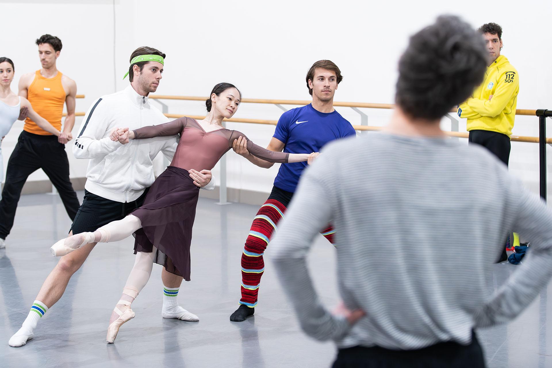 In the rehearsal room a dancer en point, one foot raised, leaning back and supported by another dancer holding her by the waist and holding a hand