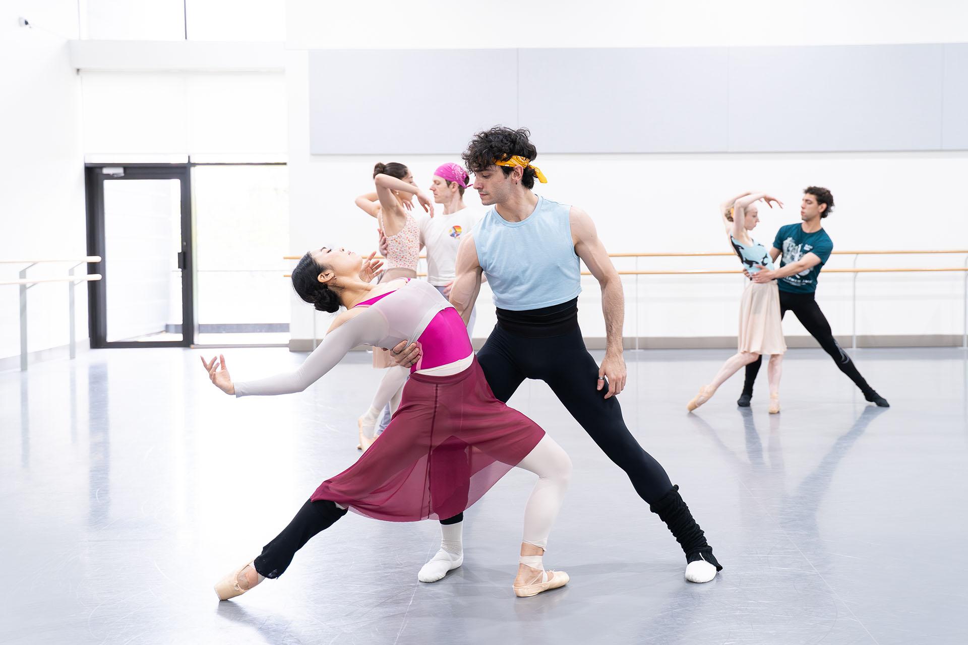 Dancer with white leggings, burgundy skirt and shiny pink leotard leans back gracefully, supported by the waist by male dancer while gazing at her