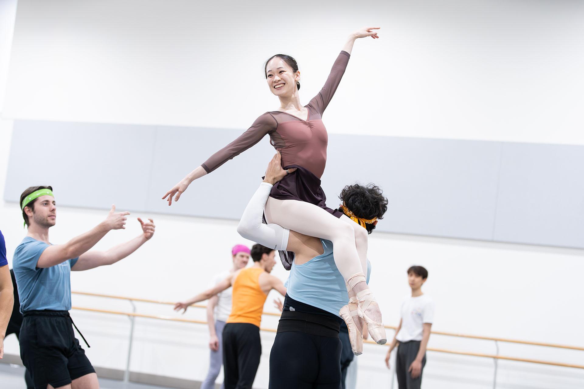 Dancer smiling as she is lifted, sat on the left shoulder of another dancer, one arm angled up, the other angled low