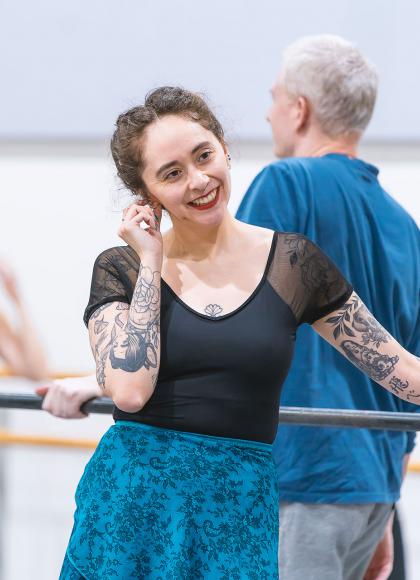 Woman in black top and blue slip stands at the barre, smiling