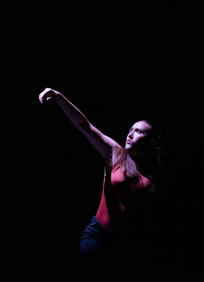 Woman against a black background wearing red, one arm raised, face lit and 